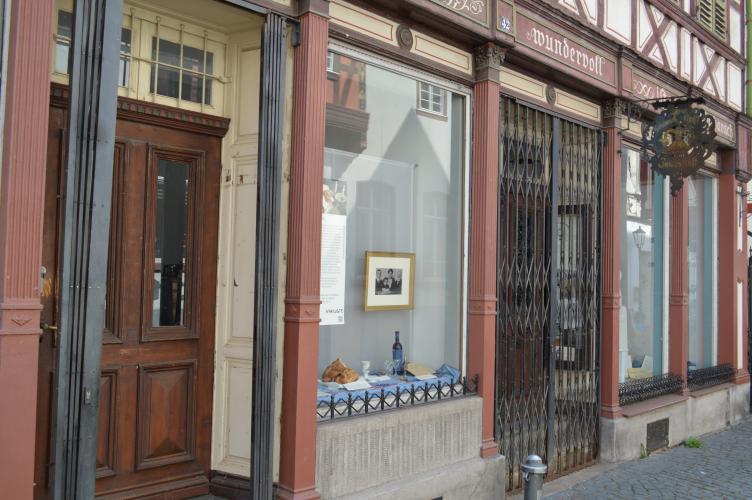 Authentisch überlieferte Schaufensterzeile mit Türanlagen, Ziehgittern, Holzrollläden und kannelierten Säulen aus Gusseisen. Die gemalte Werbebeschriftung in den Feldern über den Fenstern sowie der geschmiedete Werbeausleger über der Ladentür verleihen der Anlage Individualität und Ausdruck.